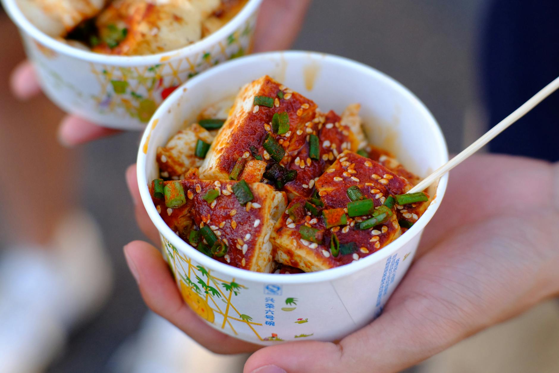 A close-up of spicy tofu with sesame and spring onions in a paper cup, held by hand.