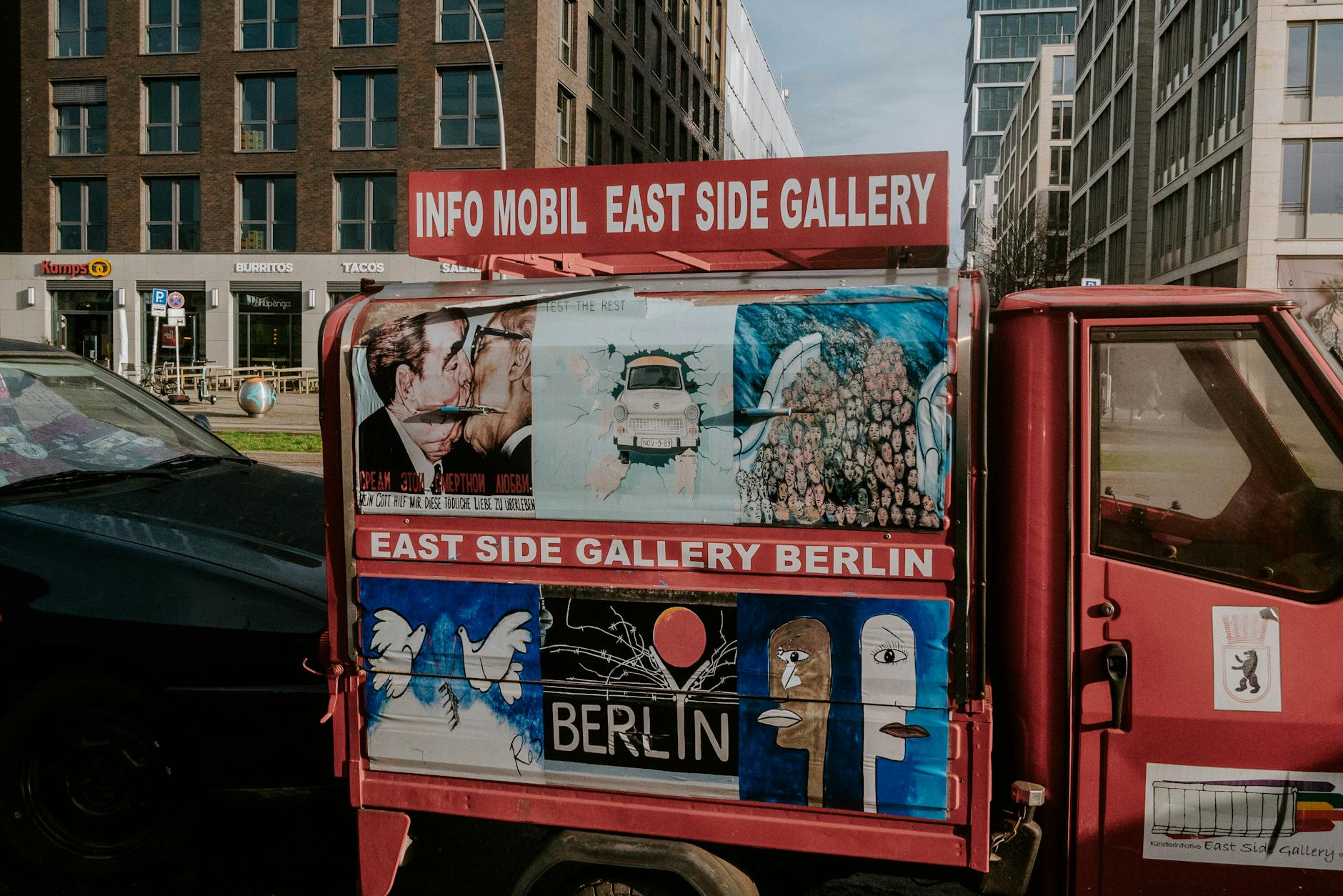 A red vehicle showcasing iconic artwork from the East Side Gallery in urban Berlin streets.
