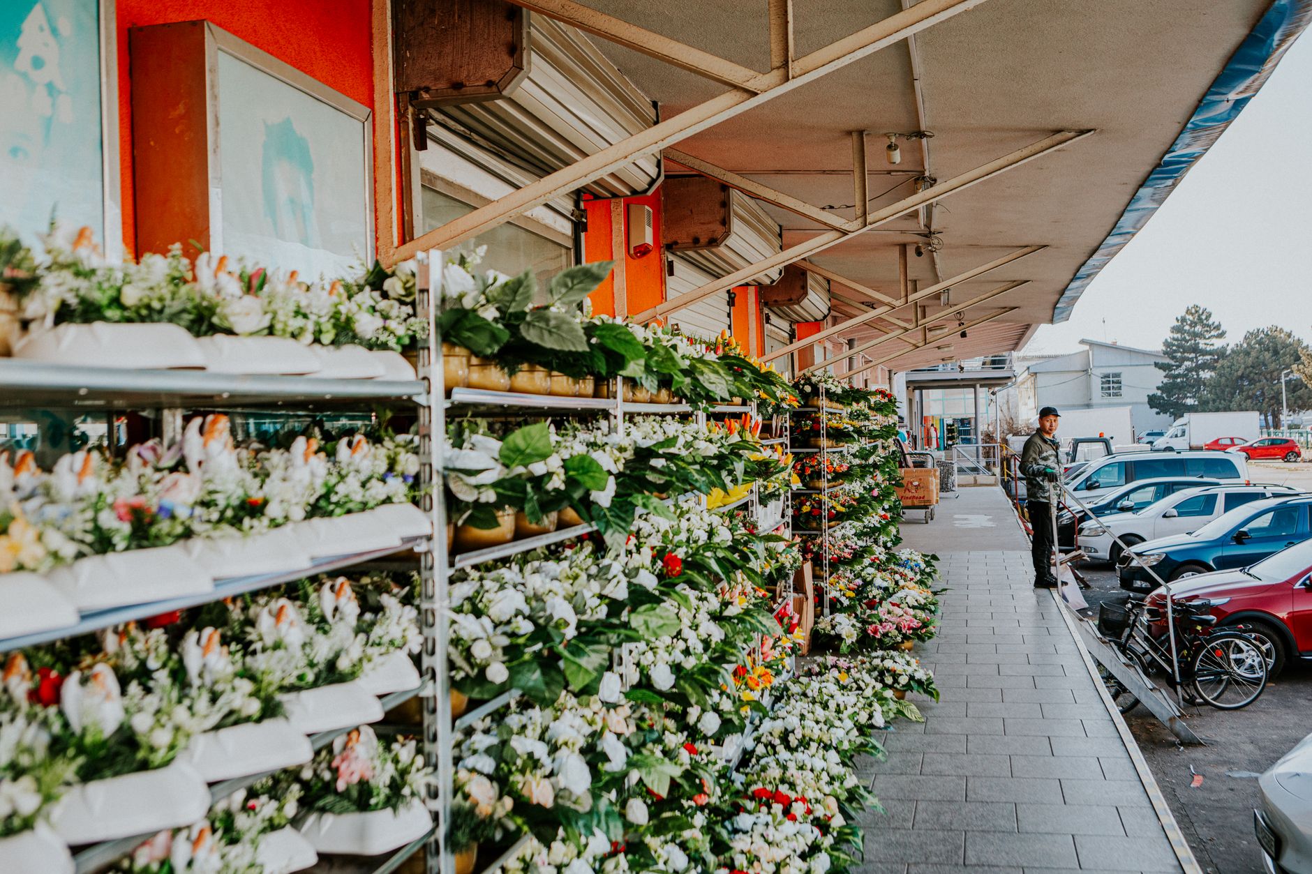 Outdoor floral market display with vibrant flowers and greenery, featuring parked cars and people.