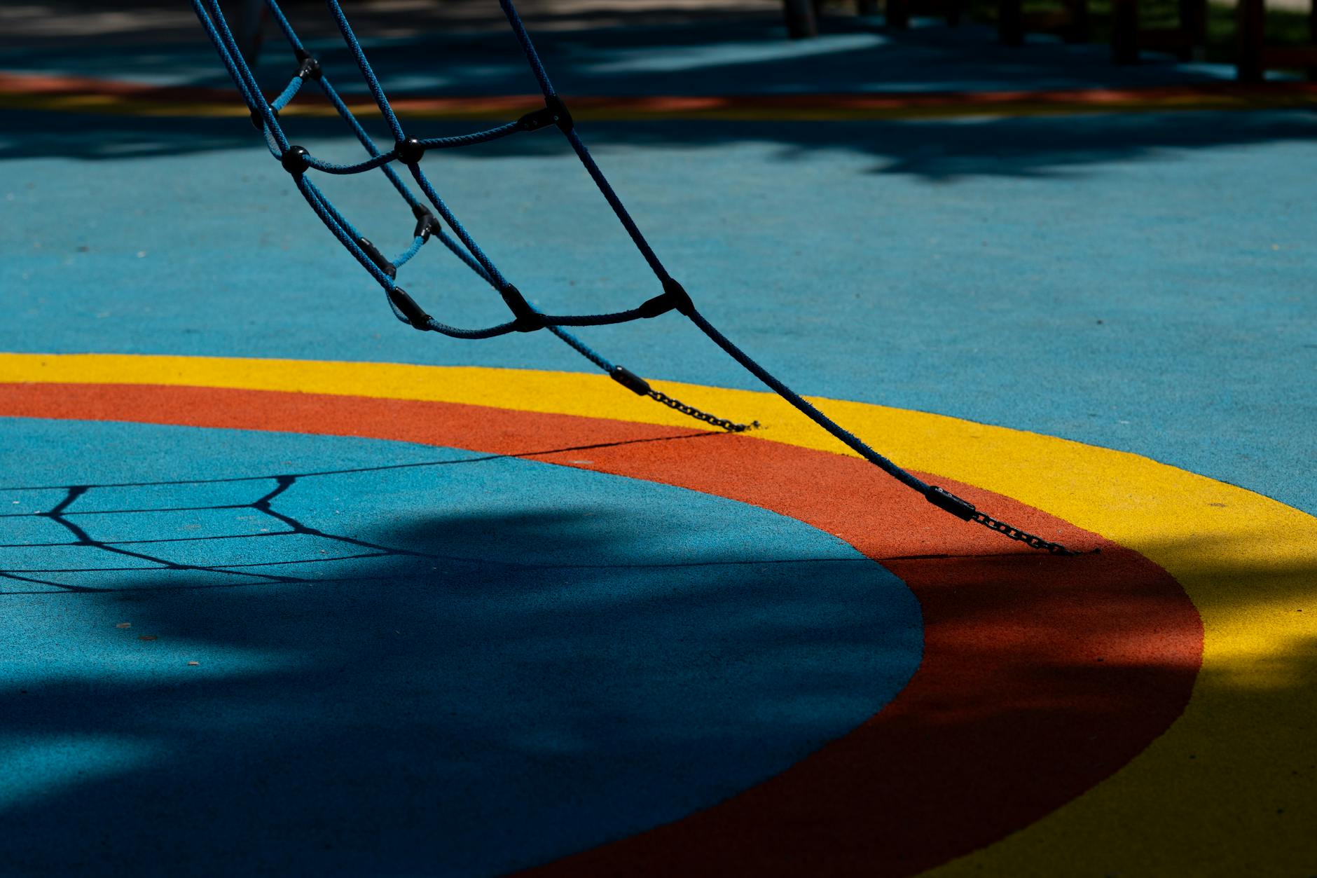 A vibrant blue, yellow, and red playground surface with rope climbing structure on a sunny day in a park.