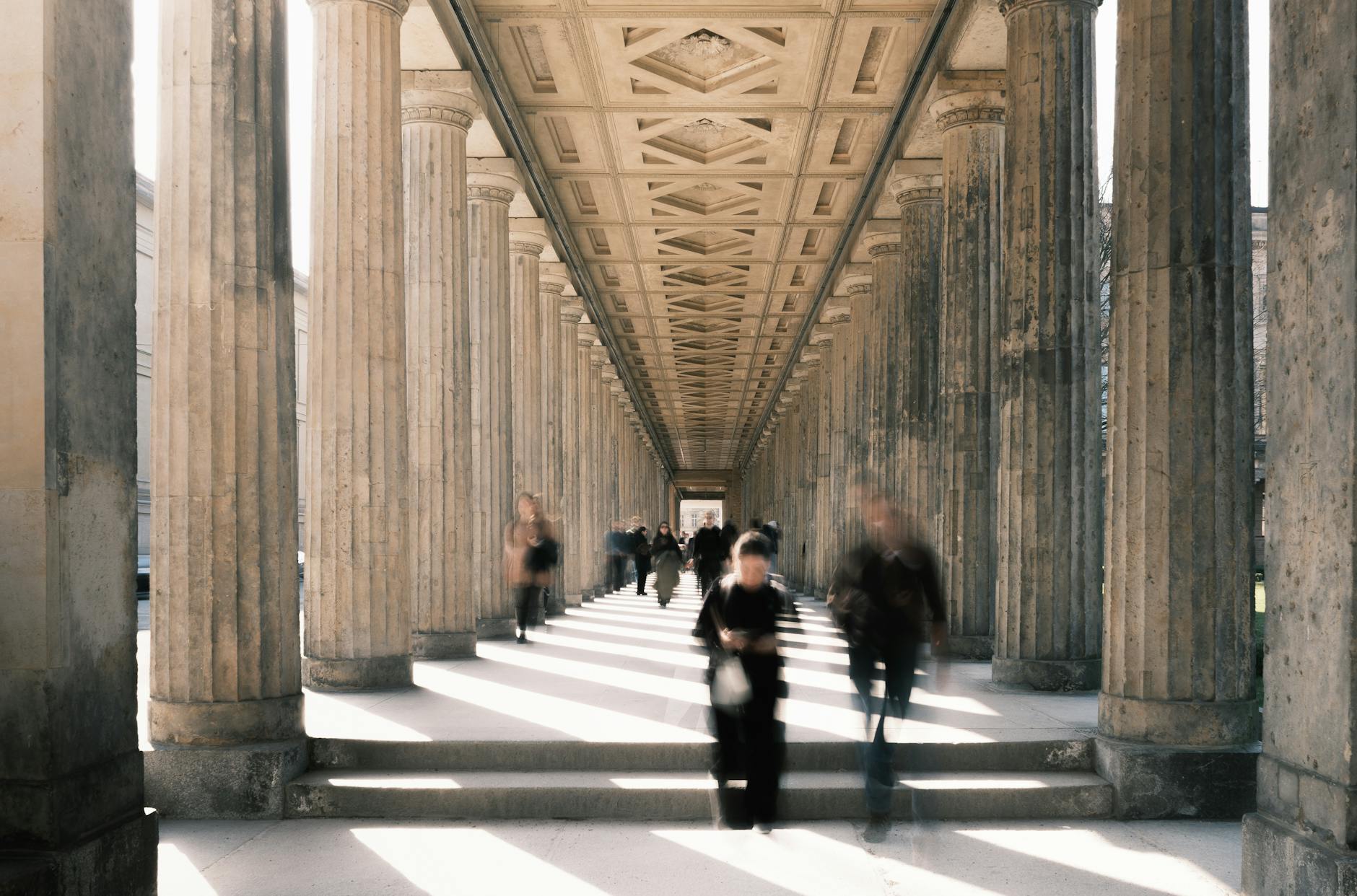 Blurred motion of people walking through a historic colonnade in Berlin, Germany, emphasizing architectural symmetry.
